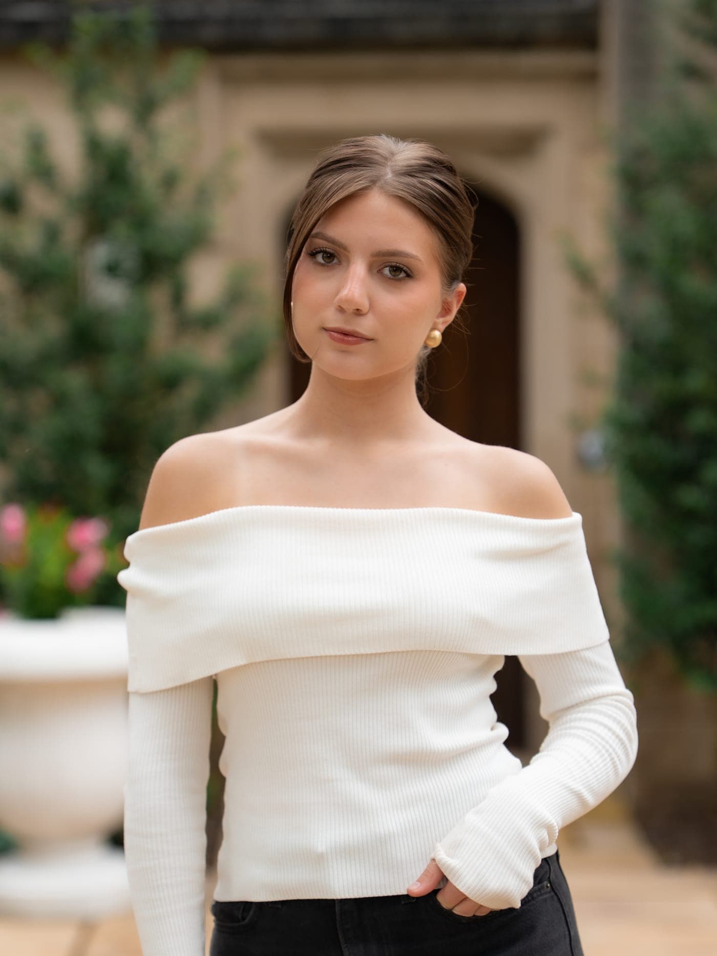 Liza standing outdoors in front of a stone building with greenery behind her, one hand in her pocket, in a cream off-the-shoulder knit and black jeans, hair swept back, wearing a soft natural look.