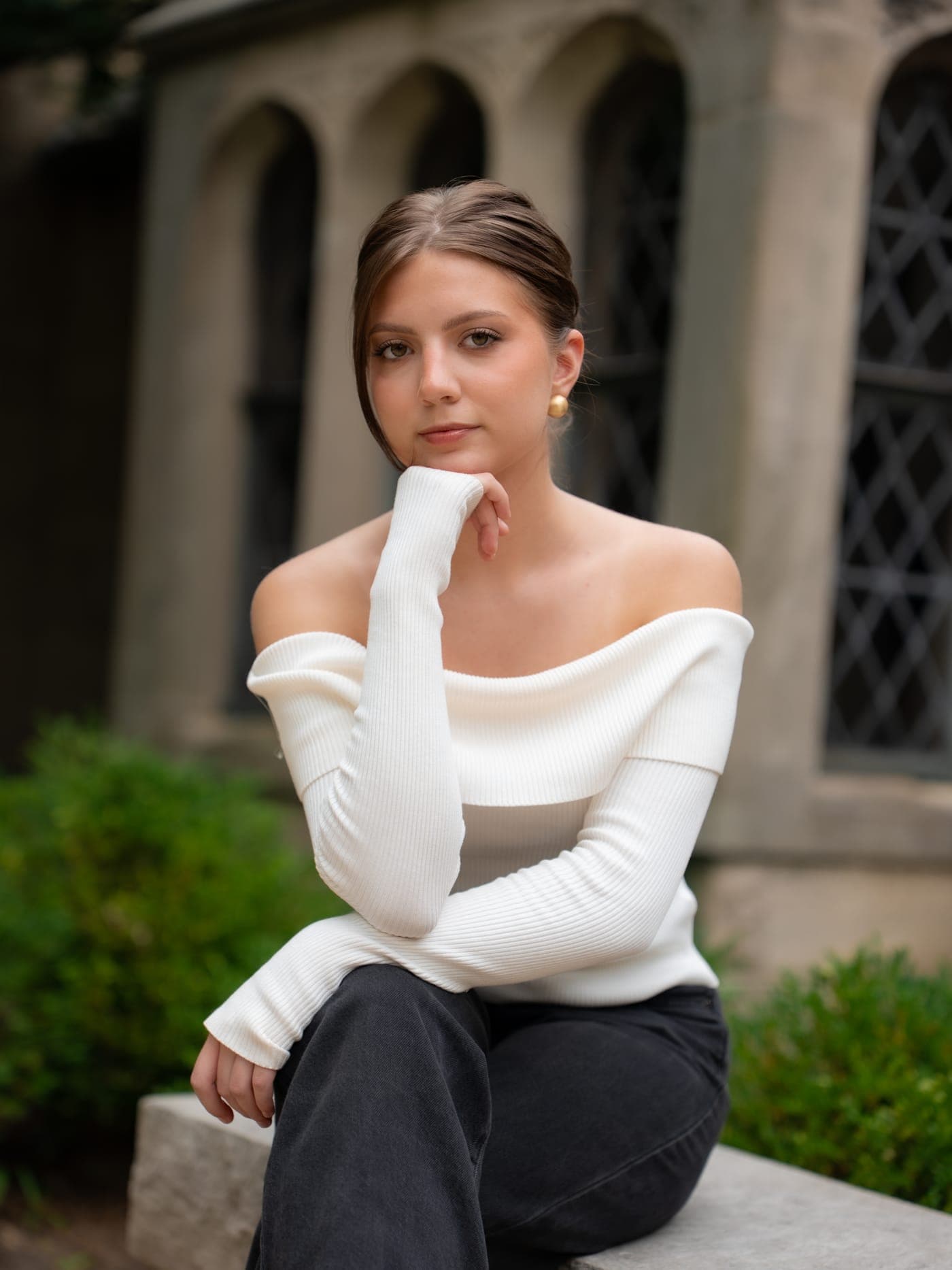 Liza seated on a stone ledge outside a stone building, chin resting on her hand, in a cream off-the-shoulder knit and black jeans, hair swept back, wearing a soft natural look.