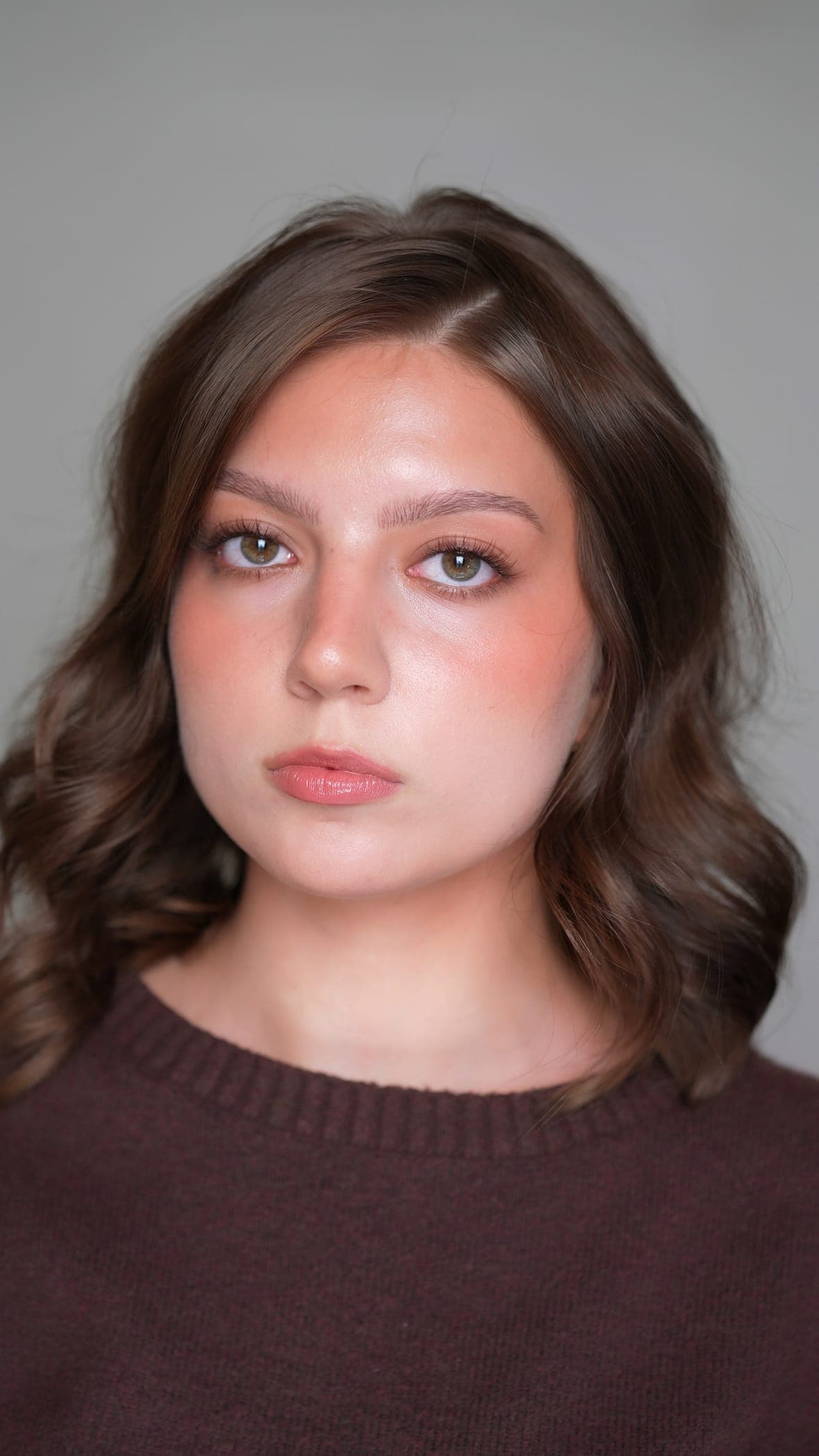 Head-and-shoulders portrait against a plain grey backdrop — shoulder-length brown hair in loose waves and a soft natural look: glowy skin, warm peach blush, a neutral eye with softly defined lashes, and a glossy rosy-nude lip.