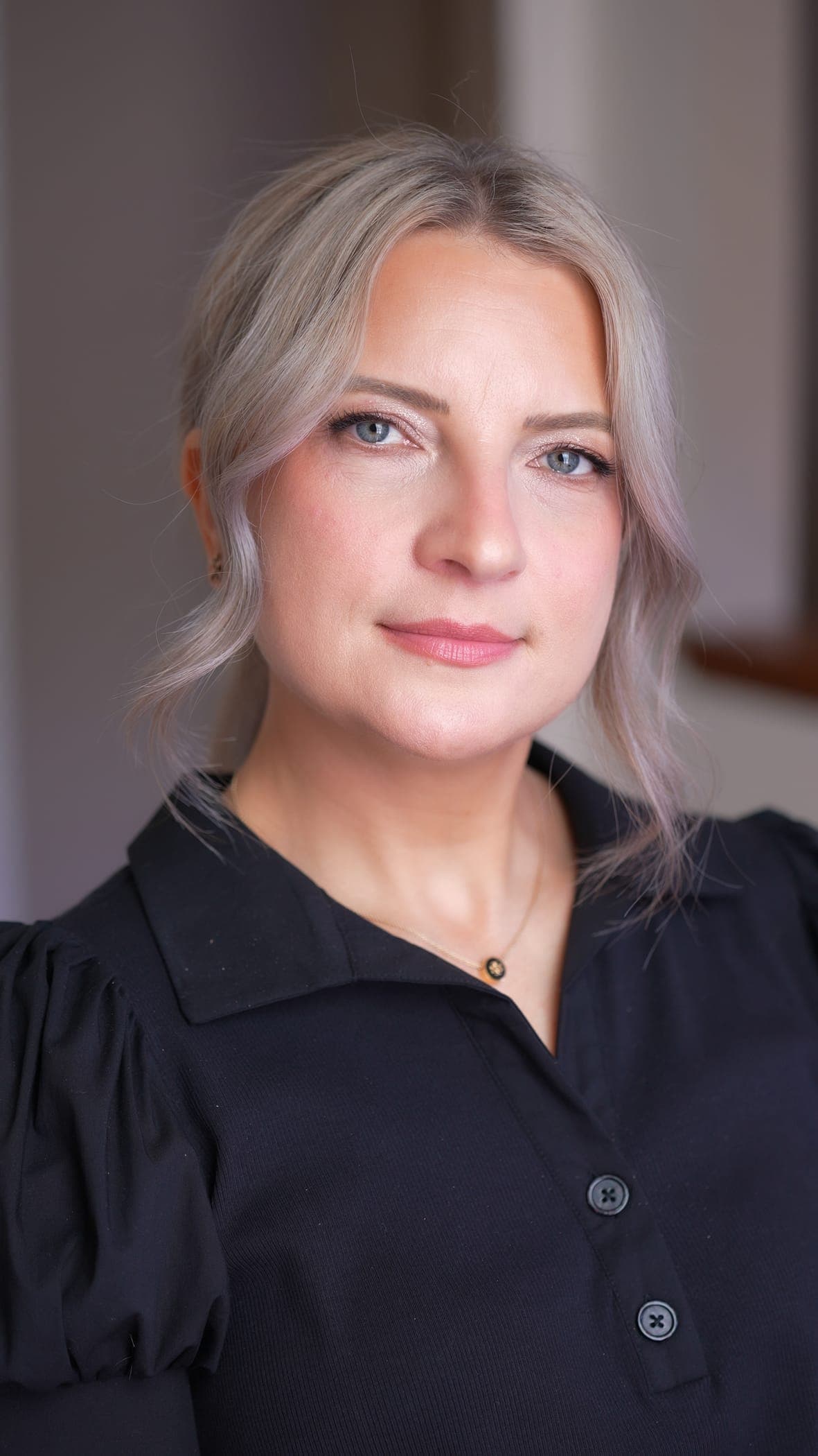 Head-and-shoulders portrait of a client with long ash-blonde waves in a black collared blouse, looking at the camera — soft glam: luminous skin, a softly shimmered neutral eye with fine liner, rosy blush, and a muted rose lip.