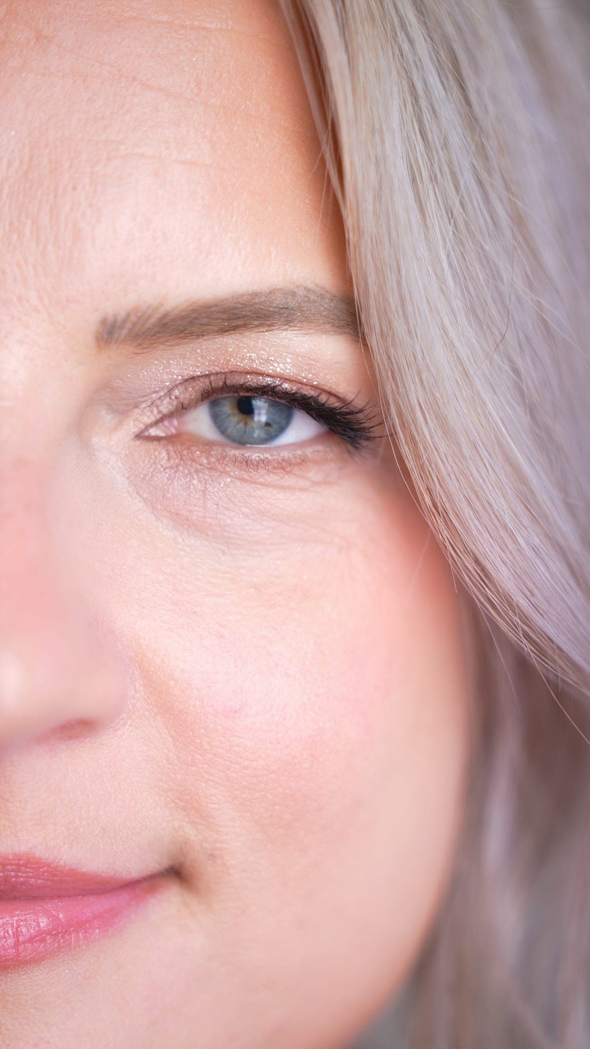 Macro detail of a client's eye — a shimmering champagne lid, fine dark liner along the upper lash line, lifted lashes, and a softly brushed brow, framed by pale blonde hair.